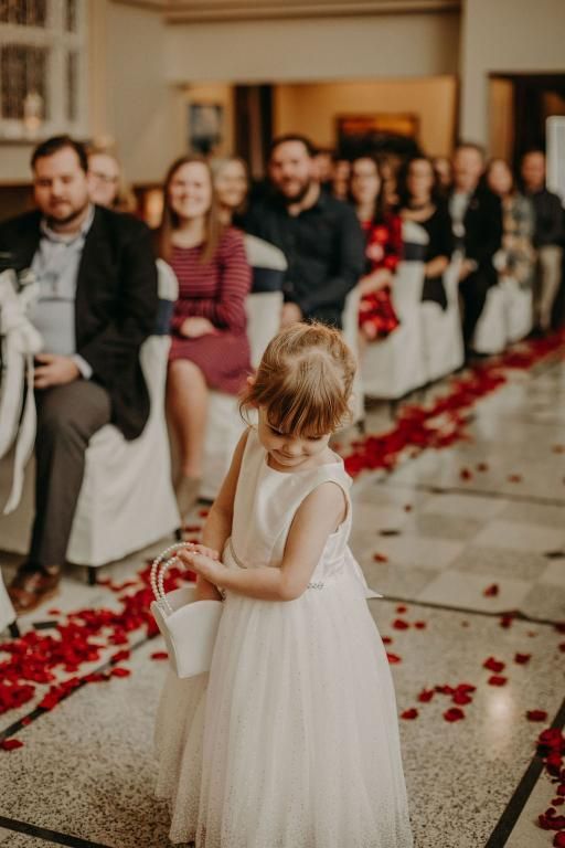 A little girl in a white dress scatters rose petals down an aisle at a wedding ceremony.
