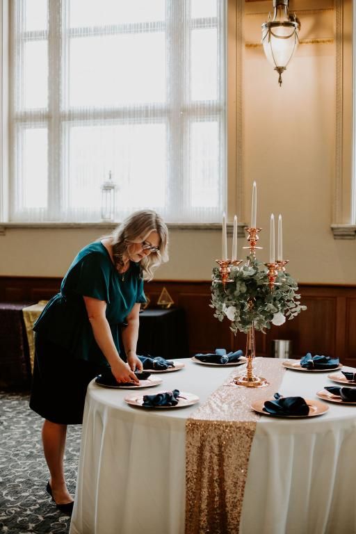 Woman setting a table with gold accents, candles, and navy napkins in a well-lit room.