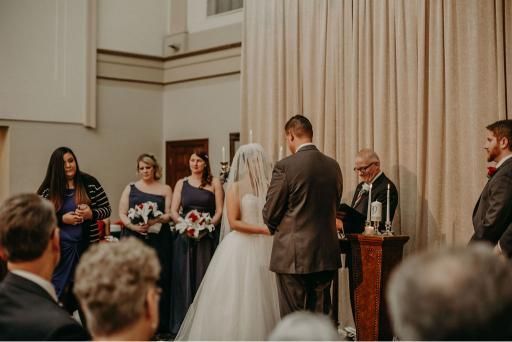 Wedding ceremony: Bride and groom at altar, bridesmaids, priest. Indoor setting with draped fabric, candles.