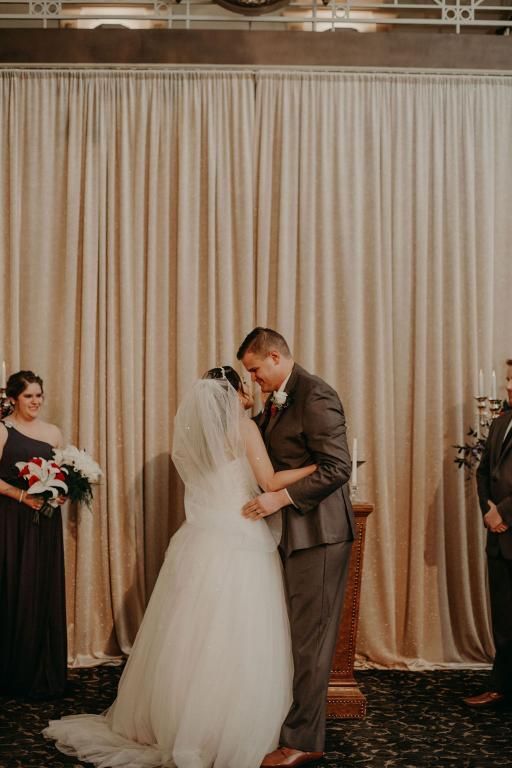 Bride and groom kissing at altar, wedding ceremony; cream curtain backdrop, bridesmaid watches.