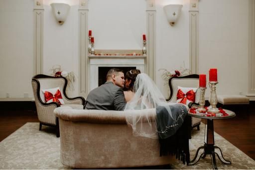 Couple kissing in a vintage bathtub, decorated with red candles and pillows, in a formal room with a fireplace.