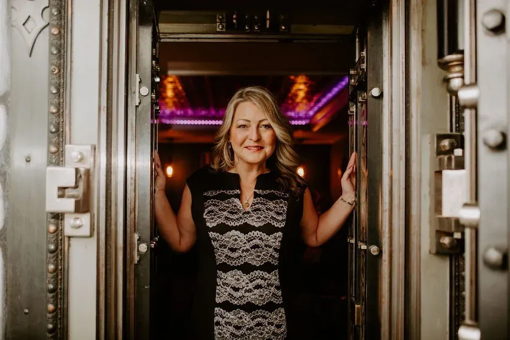 Woman in a black and white dress stands in the doorway of a bank vault, smiling.