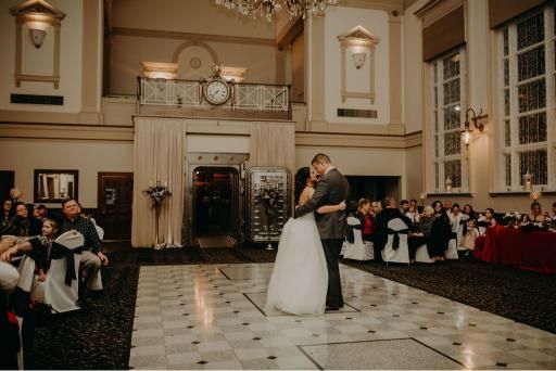 Bride and groom dance at a wedding reception in a grand hall with guests watching.