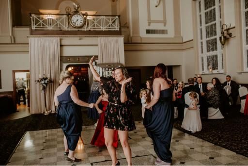 People dancing at a wedding reception. Woman in floral dress takes selfie, two women in blue dresses dance. Ballroom setting.