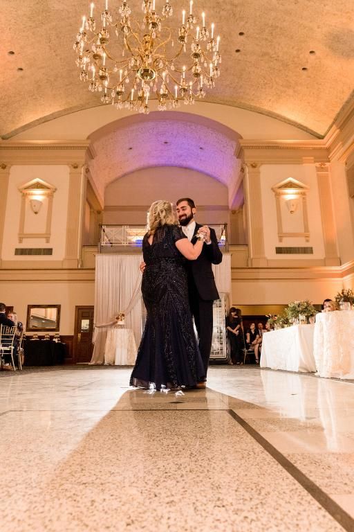 Mother and groom dancing in a ballroom with chandelier. Bride wearing a navy dress.