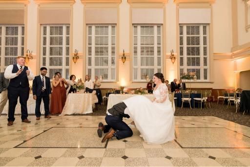 Groom kneels under bride's dress on dance floor, guests laugh. Wedding reception in ornate room.