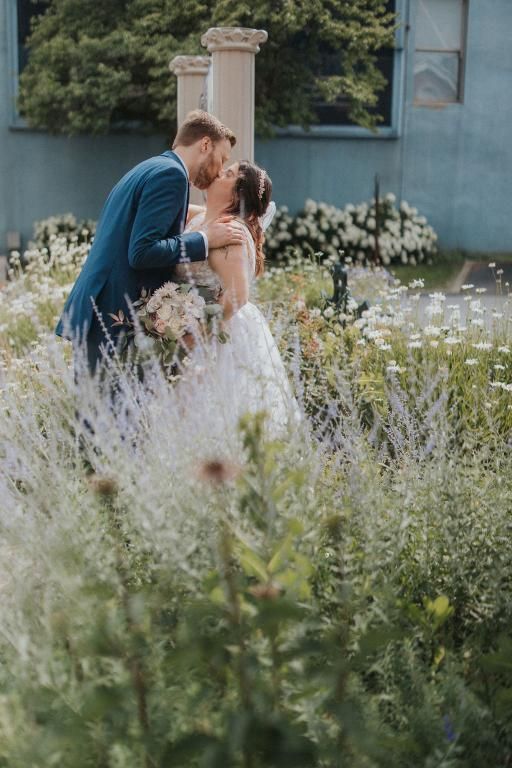 Bride and groom kissing in a garden; groom in blue suit, bride in white gown, surrounded by flowers.