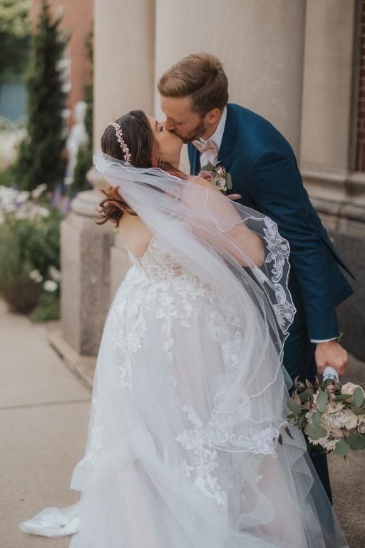 Bride and groom kissing outdoors. The bride wears a white gown and veil; the groom, a blue suit.
