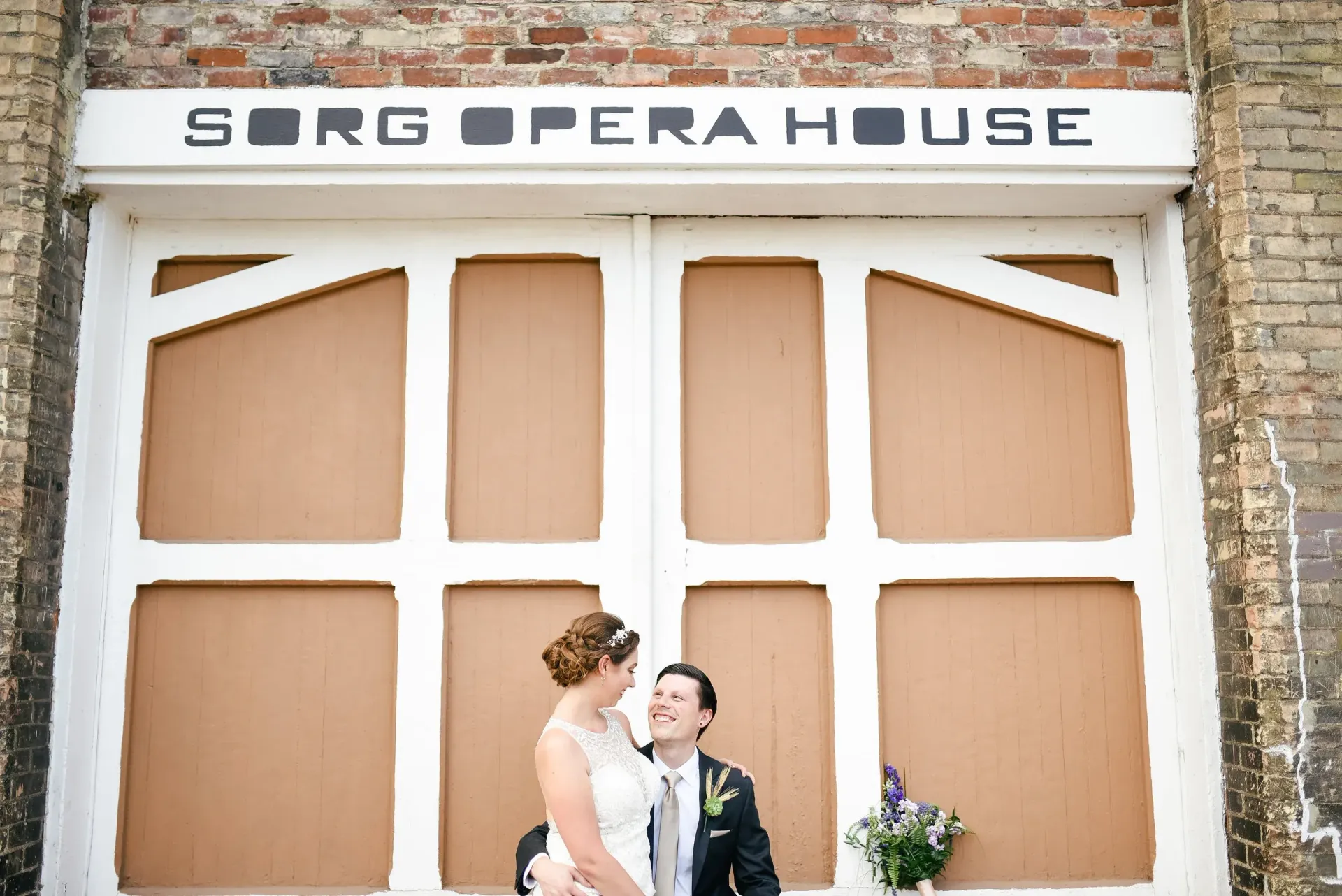 A newlywed couple smiles at each other in front of the Sorg Opera House doors. The doors are beige with black lettering.