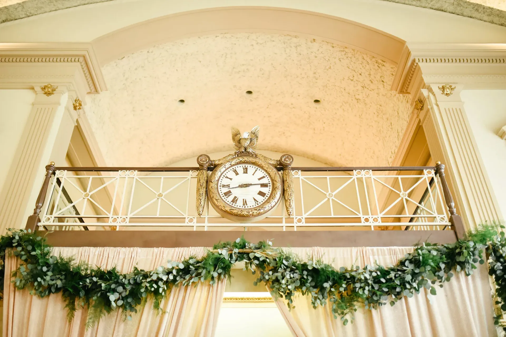 A vintage clock mounted on a balcony, decorated with greenery and draping curtains in a grand hall.