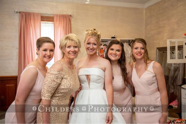 Bride poses with four women in a room, all smiling. They wear dresses in shades of pink and gold.