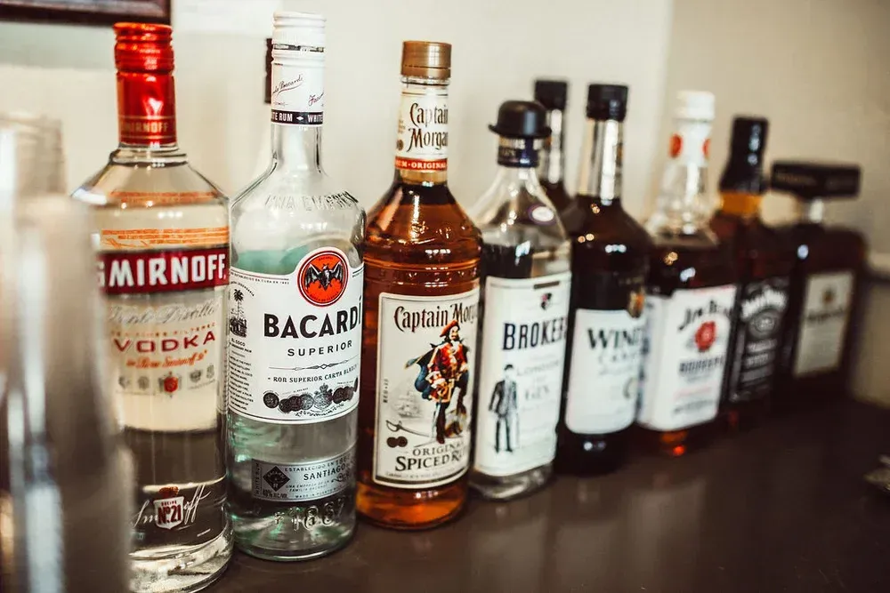 Row of various liquor bottles on a bar, including vodka, rum, and whiskey; indoor setting.