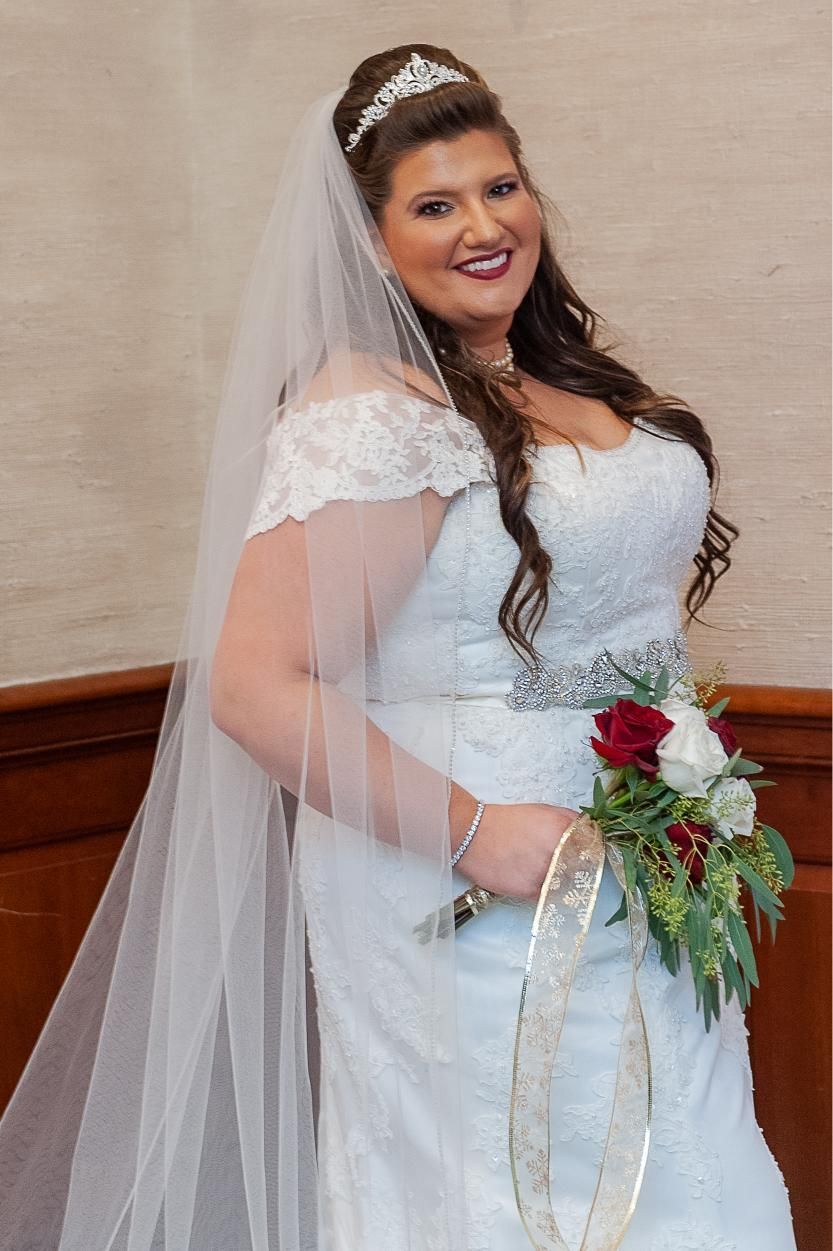 Bride in white lace wedding dress, smiling, holding bouquet, wearing veil and tiara.