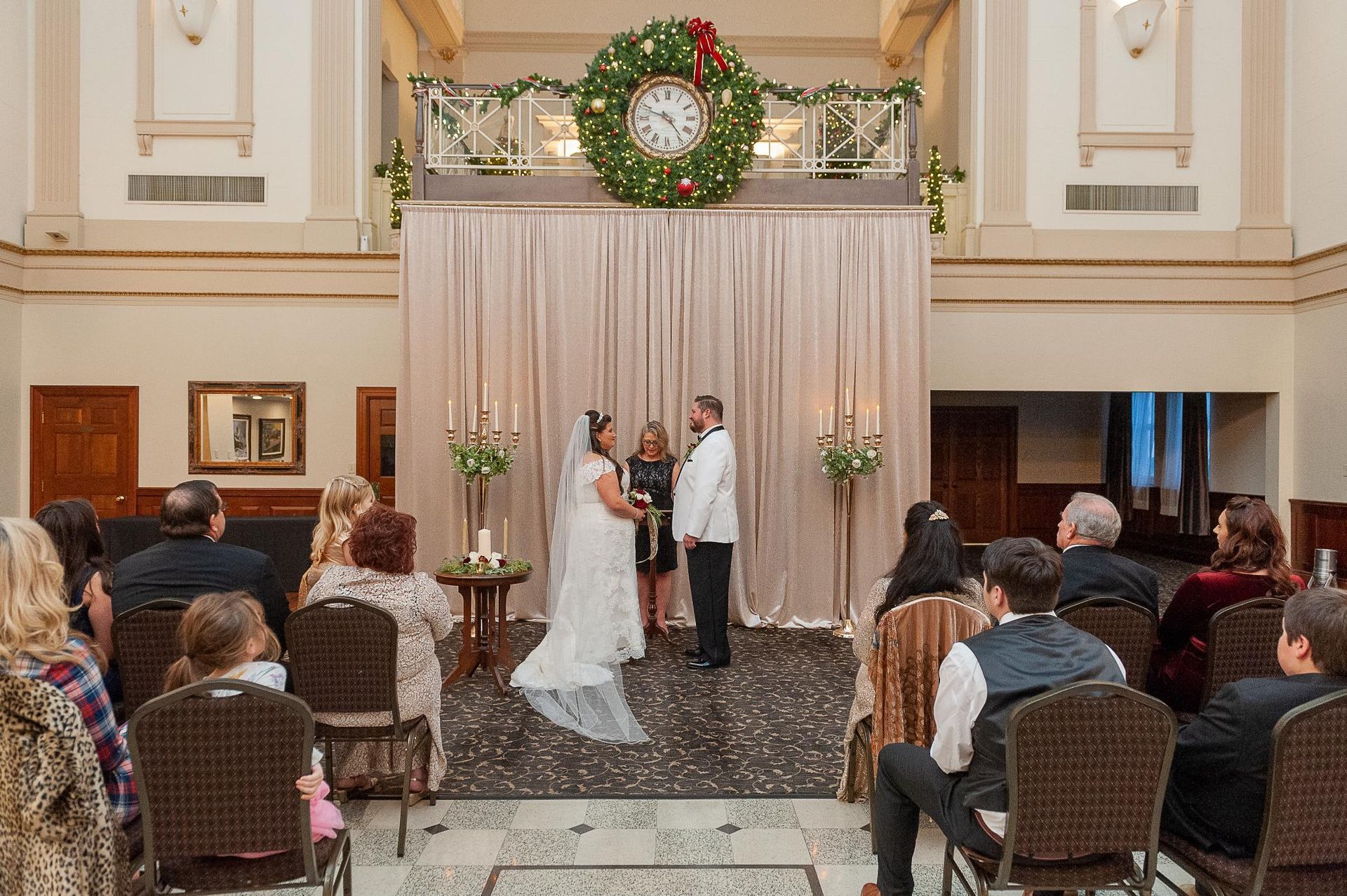 Wedding ceremony: couple exchanging vows, surrounded by guests, festive decorations in an ornate venue.