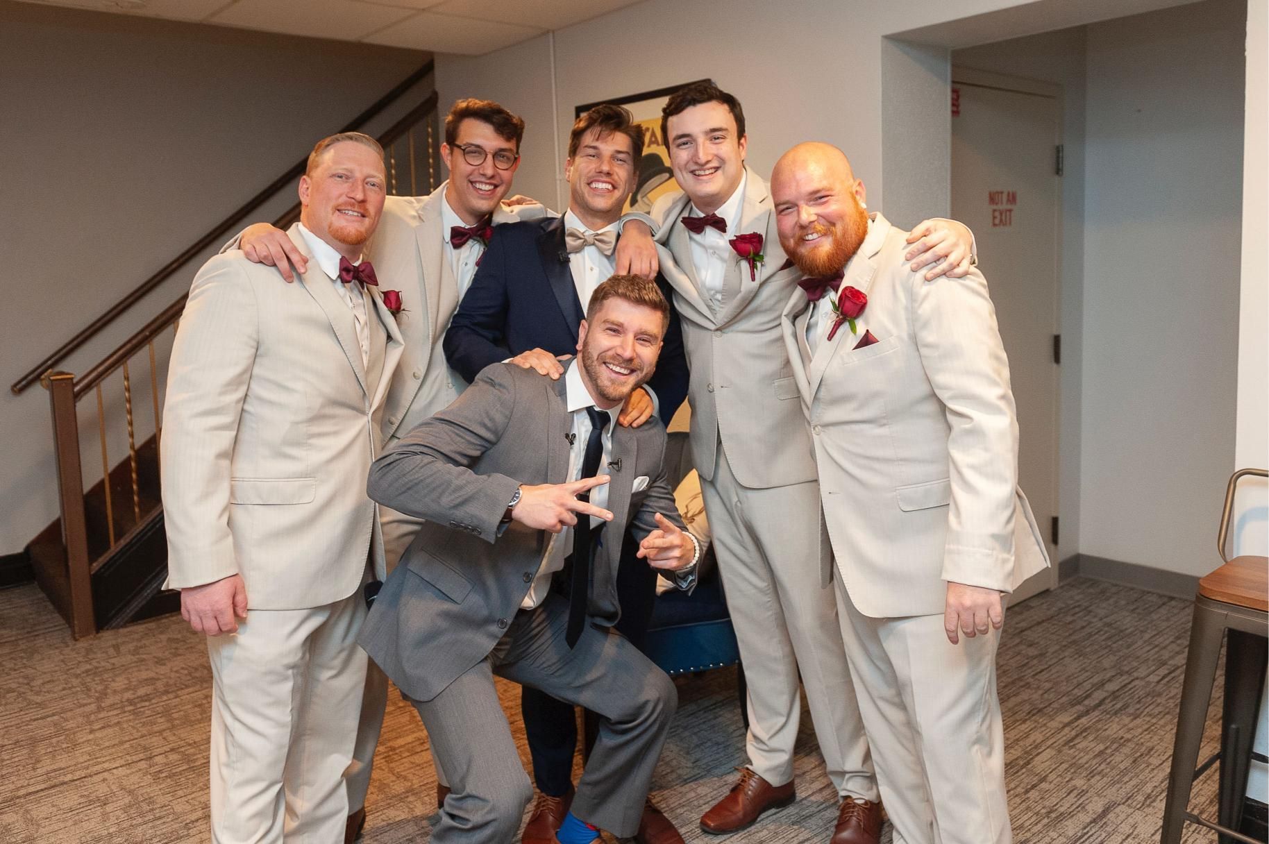 Groomsmen in suits pose for a photo in a hallway. One man is kneeling, making a peace sign.