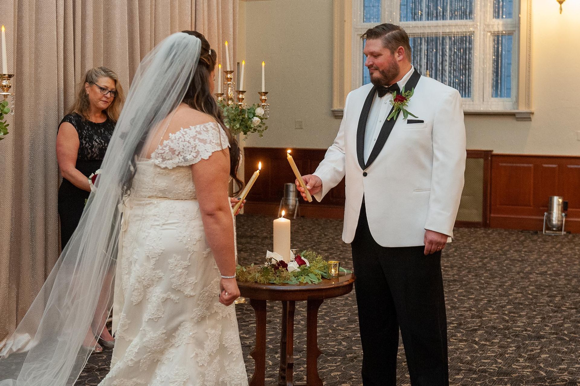 Bride and groom lighting unity candle during wedding ceremony. Woman in black dress stands by them.