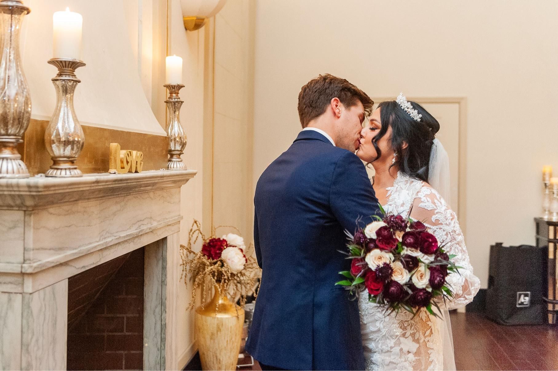 Couple kissing after wedding ceremony in a room with a fireplace, candles, and flowers.