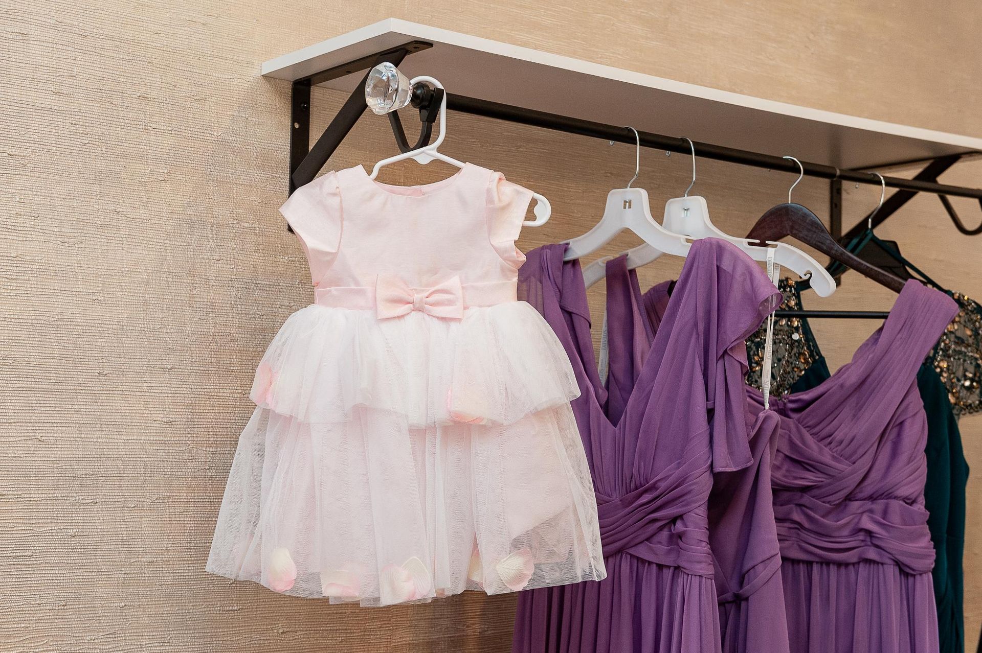A light pink toddler dress and purple bridesmaid dresses hanging on a rack.
