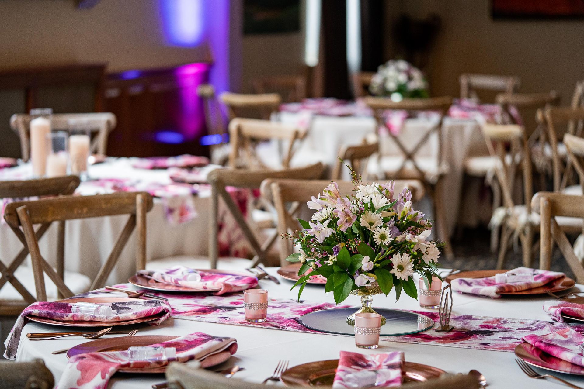 Tables set for a wedding reception. Floral centerpieces, pink floral tablecloths, wooden chairs.