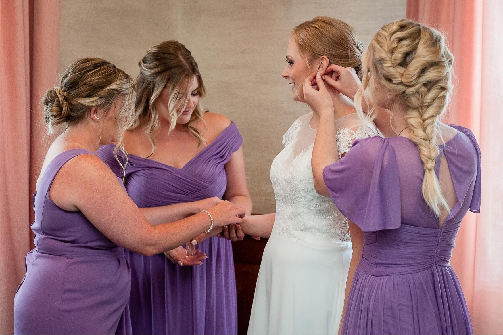Bride with bridesmaids: Bride in white dress, bridesmaids in purple helping with jewelry in a room.