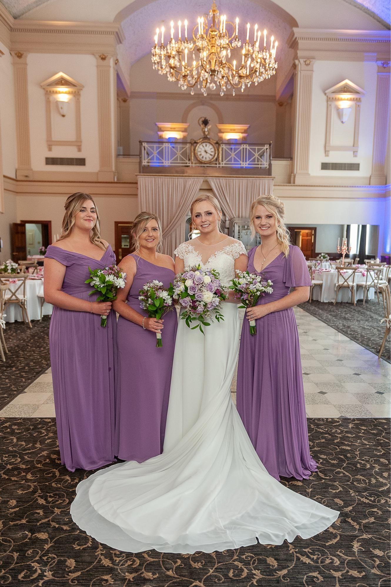 Bride with bridesmaids in a formal hall; bride in white gown, bridesmaids in purple dresses, holding bouquets.