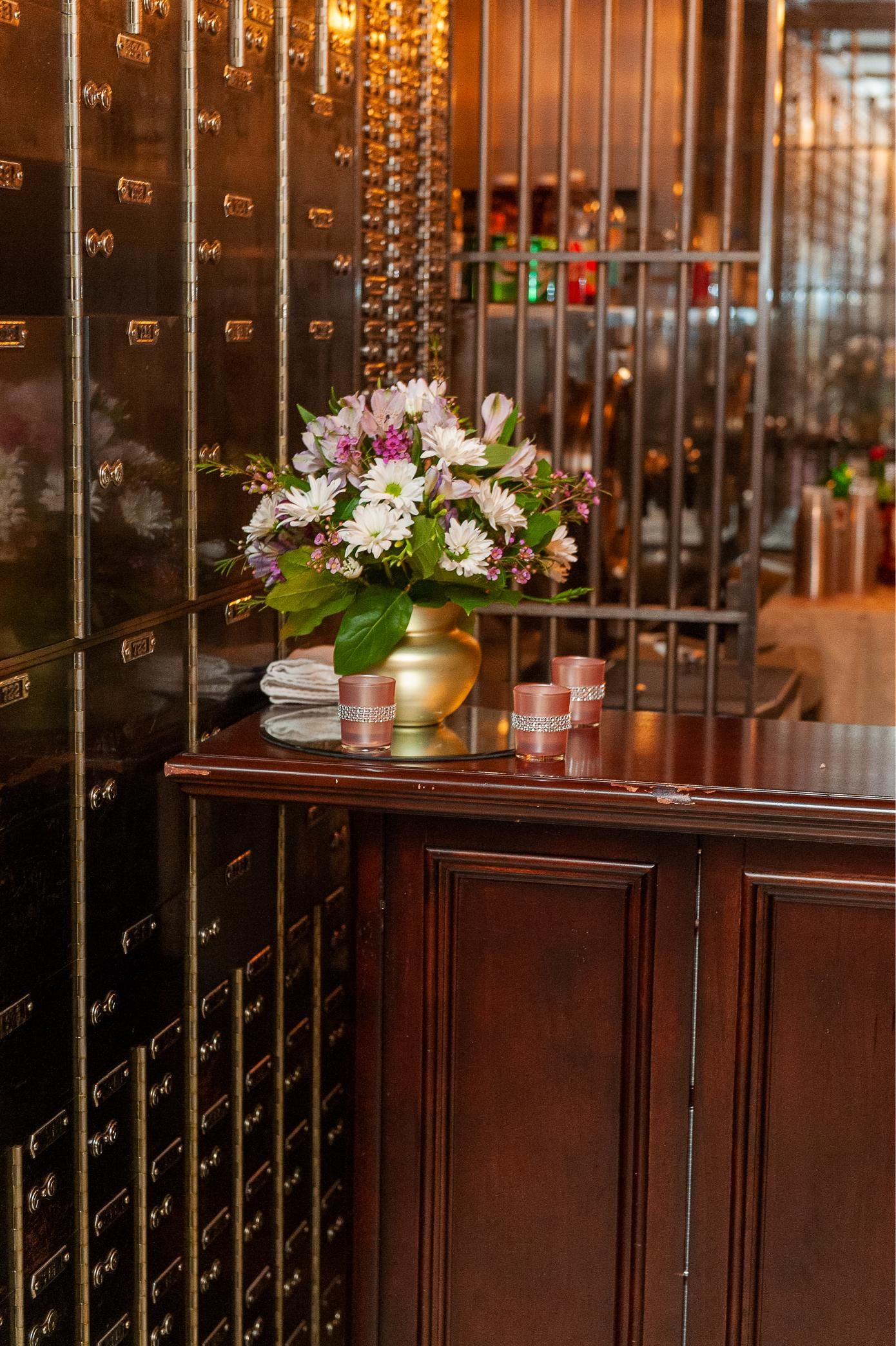 Flowers in gold vase on a dark wood counter in front of a bank vault.