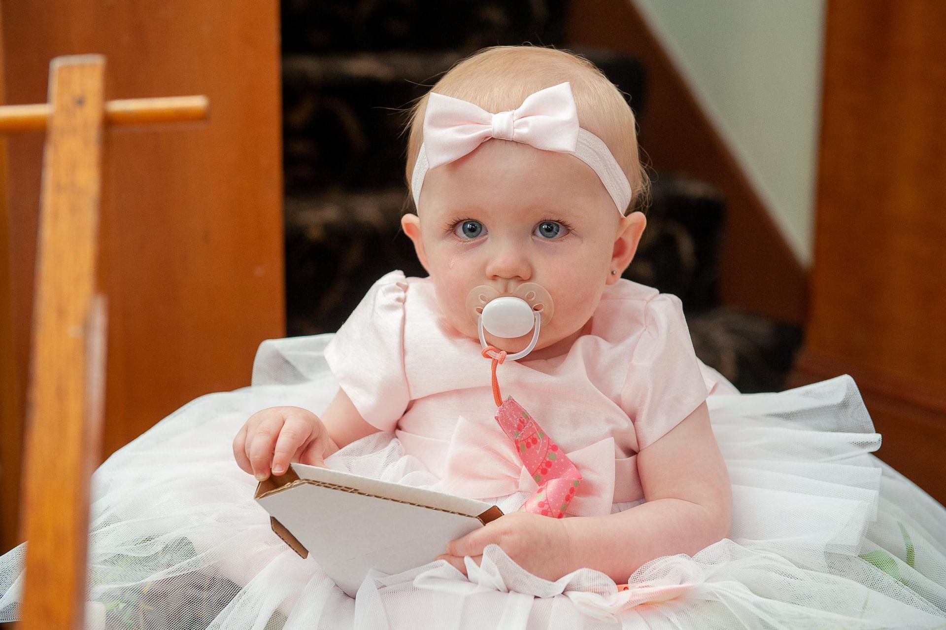 Baby in pink dress and bow, holding a card, with pacifier, sitting on stairs.