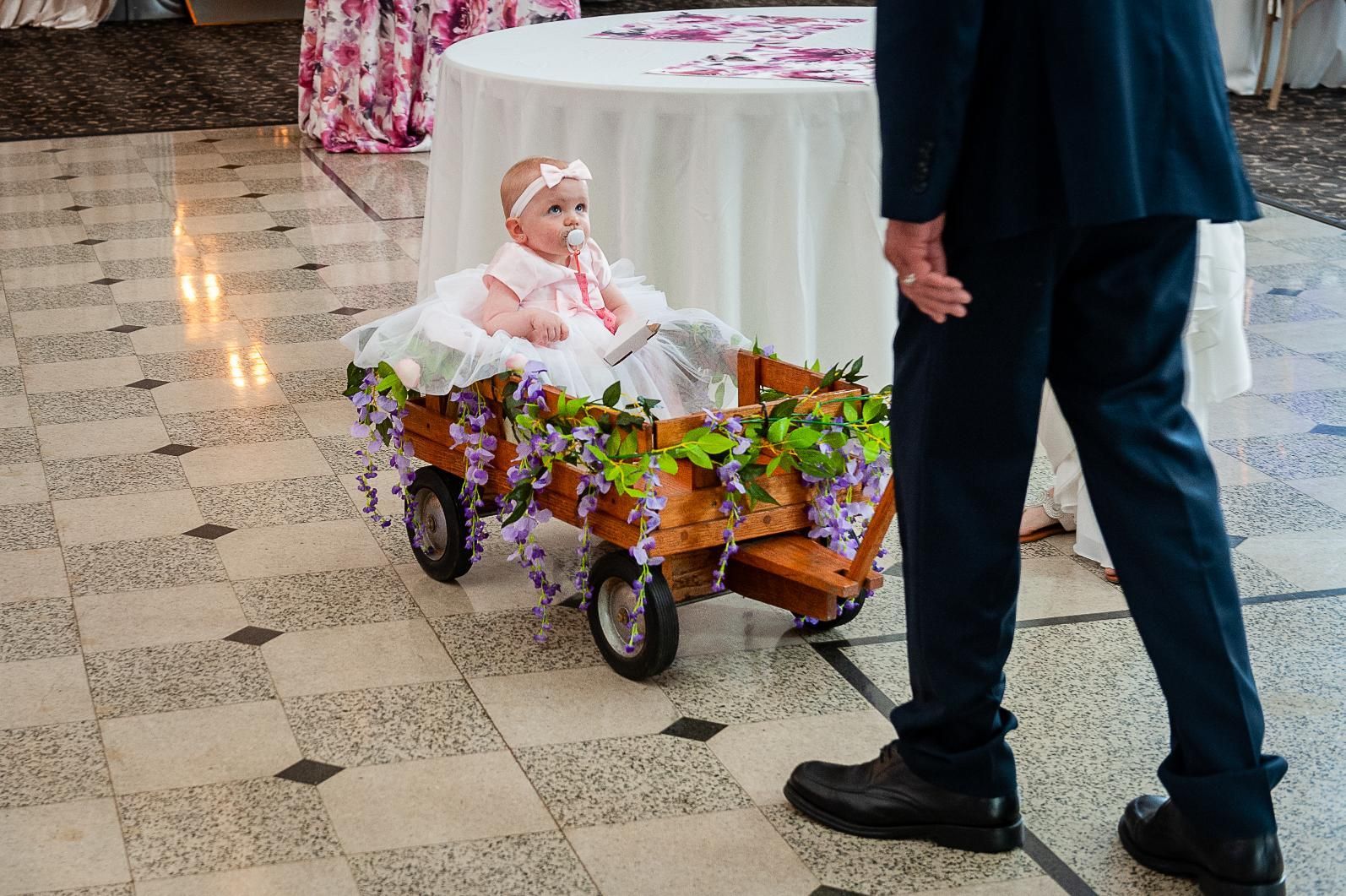 A baby sits in a decorated wooden wagon, pulled by a person in a suit, indoors on a checkered floor.