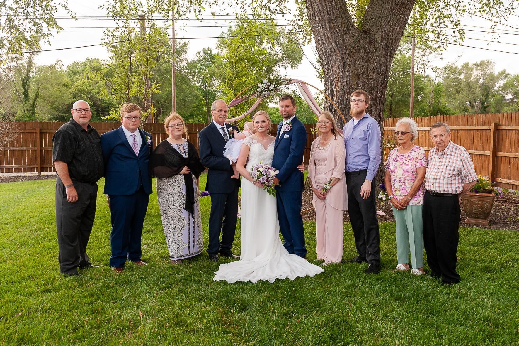 A wedding party poses outdoors on a lawn, including the bride and groom, with family.