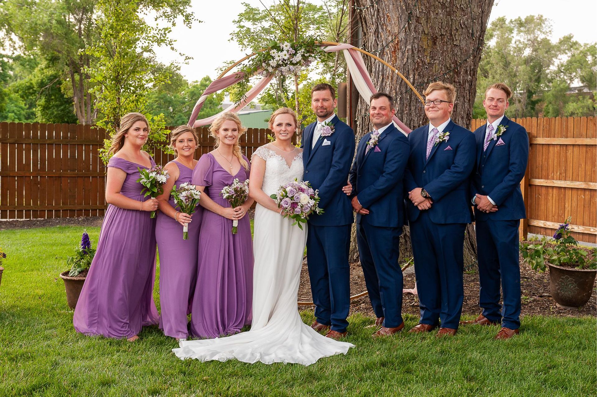 Wedding party poses outdoors: bride in white, bridesmaids in purple, groomsmen in blue, under floral arch.
