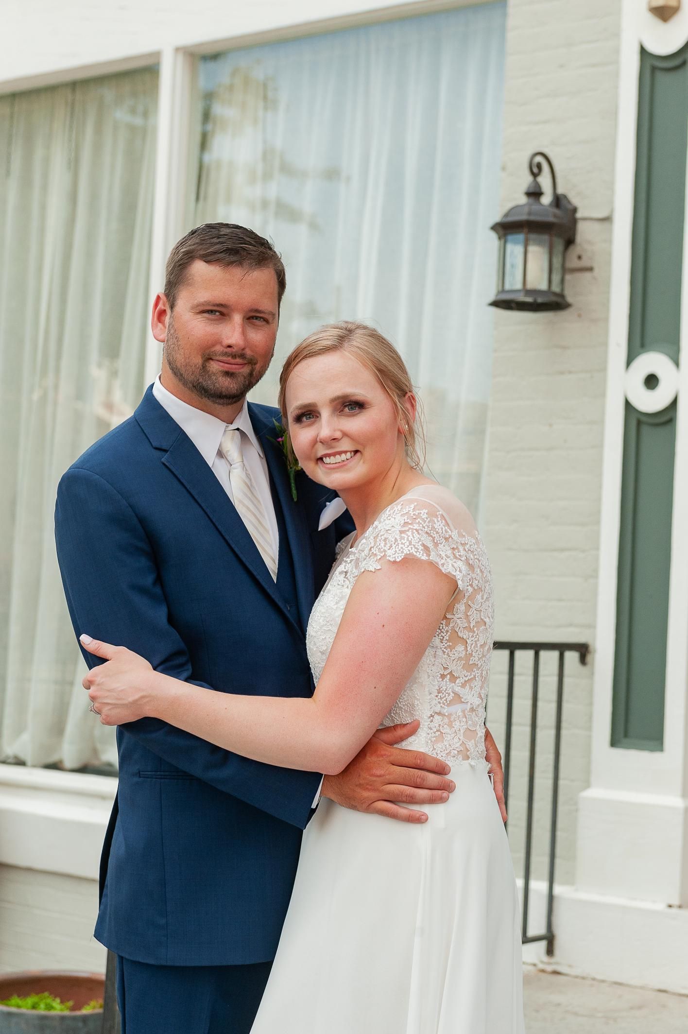 Newlywed couple embracing, smiling. Man in blue suit, woman in white wedding dress, outside building.