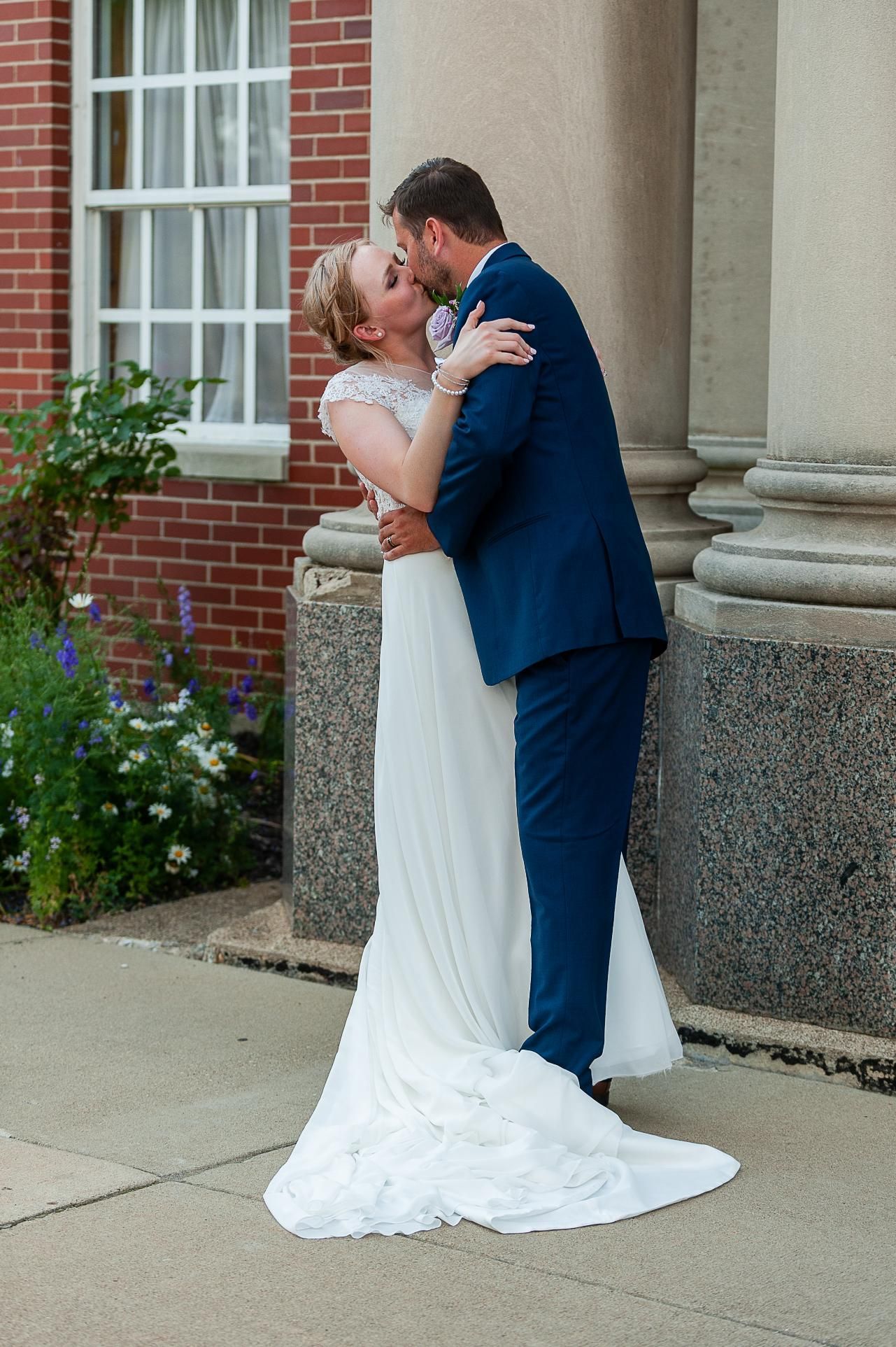 Bride and groom kissing; she's in a white dress, he's in a blue suit.  Exterior shot near brick building and columns.