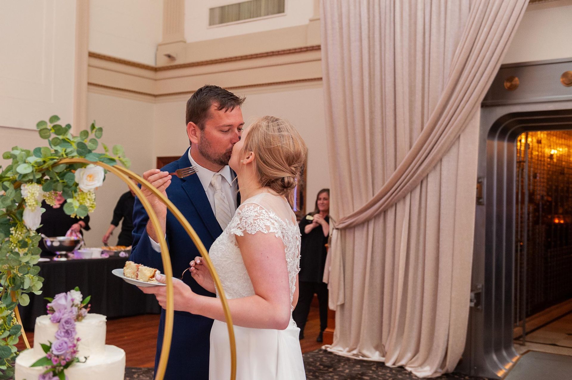 Newlyweds kiss while feeding each other cake at a wedding. A tiered cake is in the foreground.