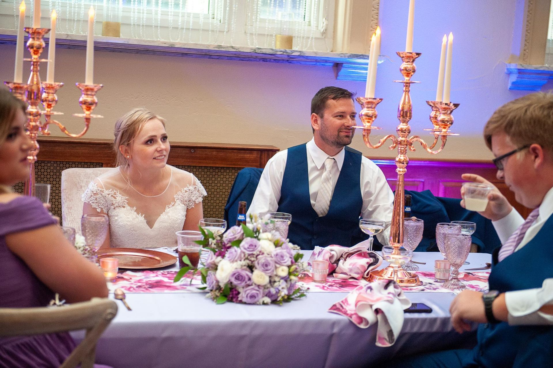 Bride and groom at wedding reception table. Bride in lace dress, groom in vest, centerpieces, guests.