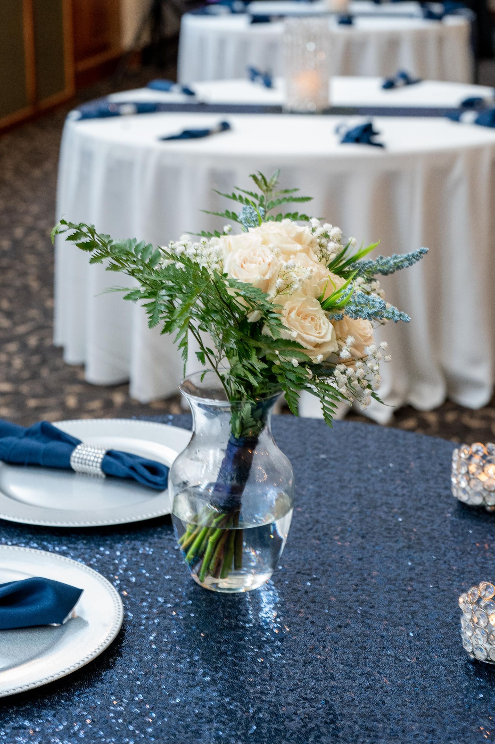 Bouquet of white roses and greenery in a vase on a navy blue sequined table, set for an event.