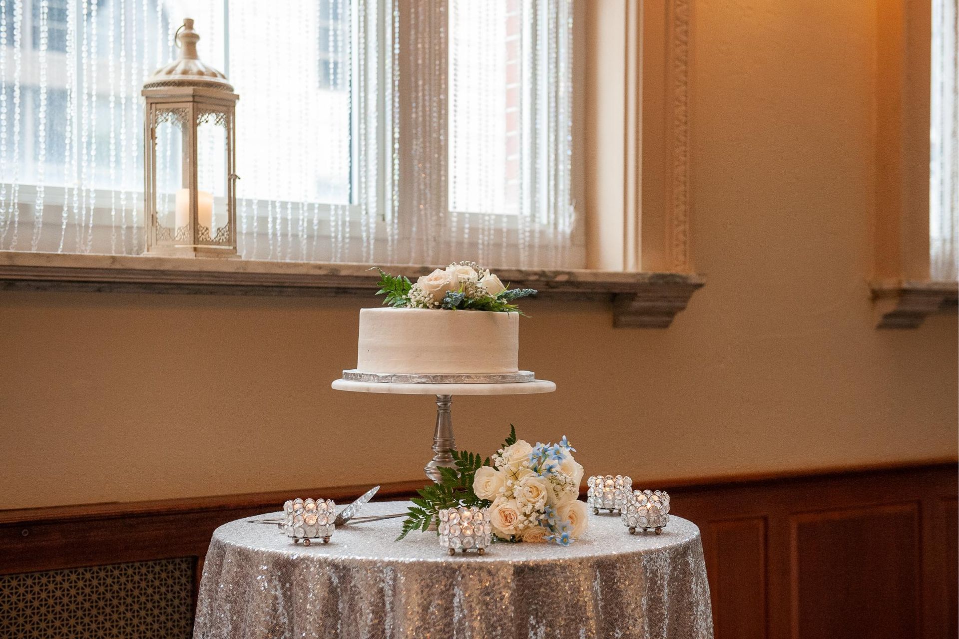 Wedding cake on a silver-covered table with flowers and candles, in front of a window.