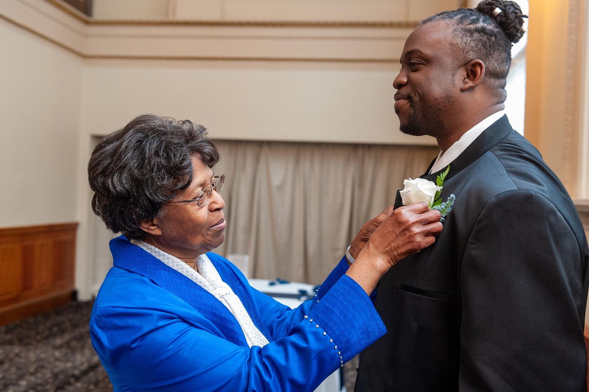 Woman in blue jacket pinning a boutonniere on a man in a black suit. Indoors.