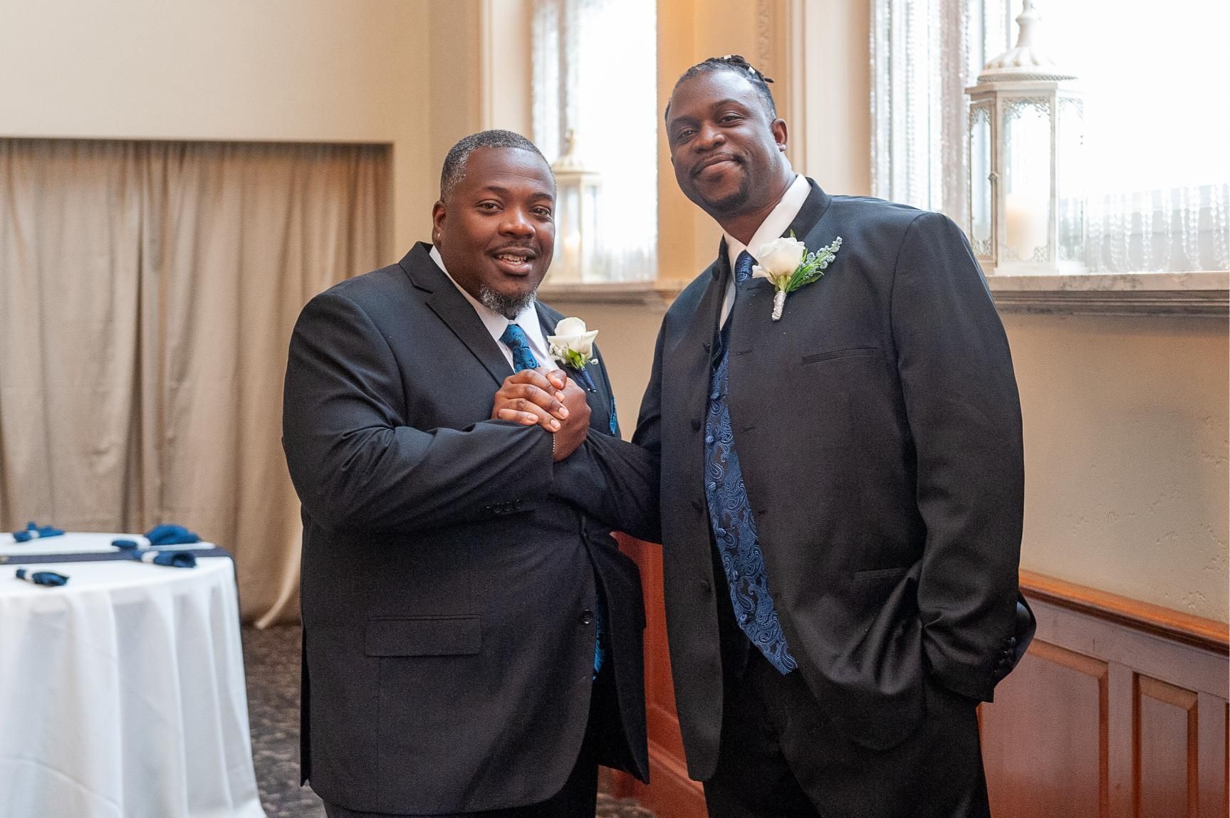 Two Black men in suits shaking hands, smiling, in a room with a window.