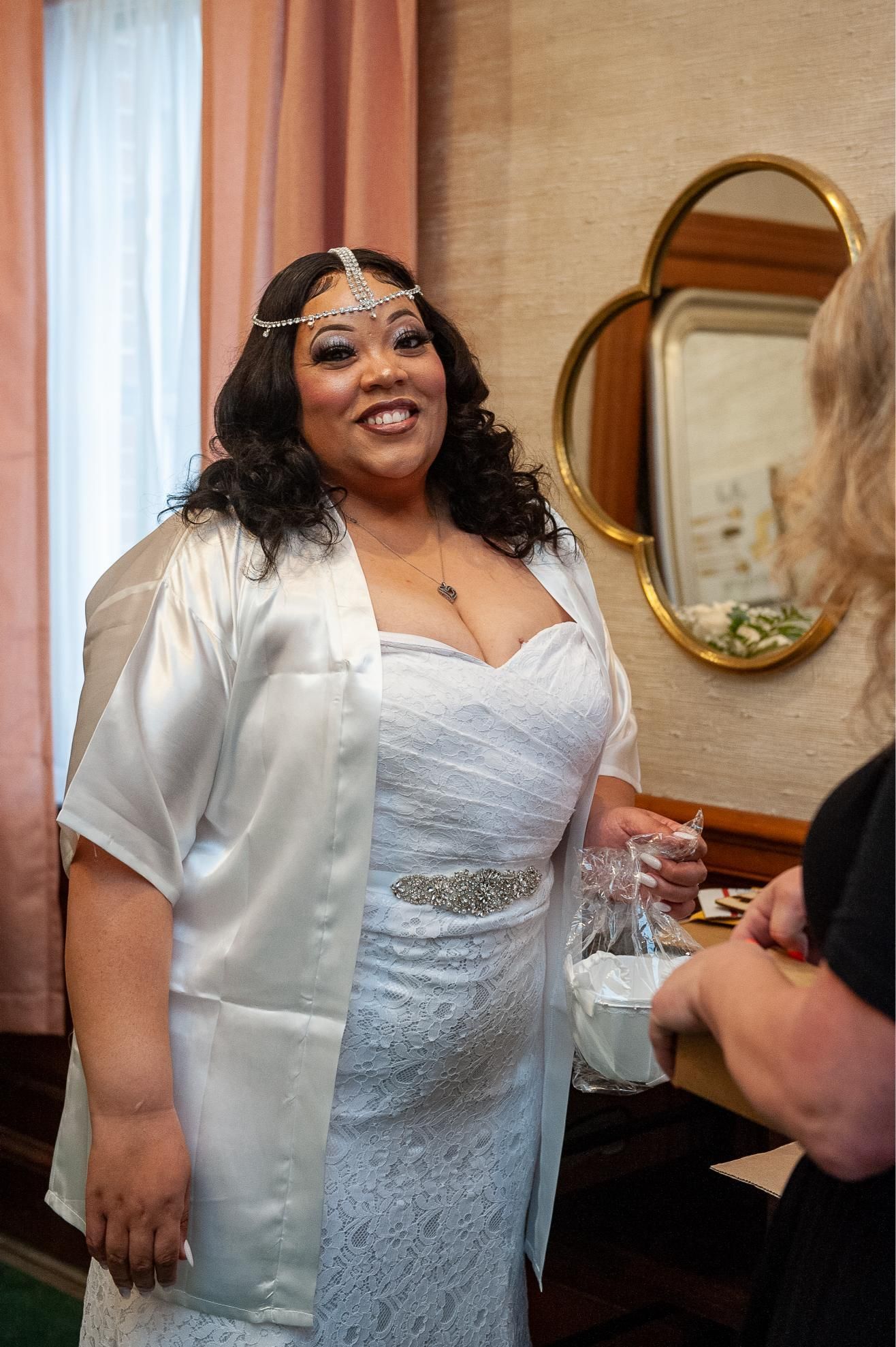 Bride in white dress smiles, holding gift bag. In a room with mirror, pink curtains.