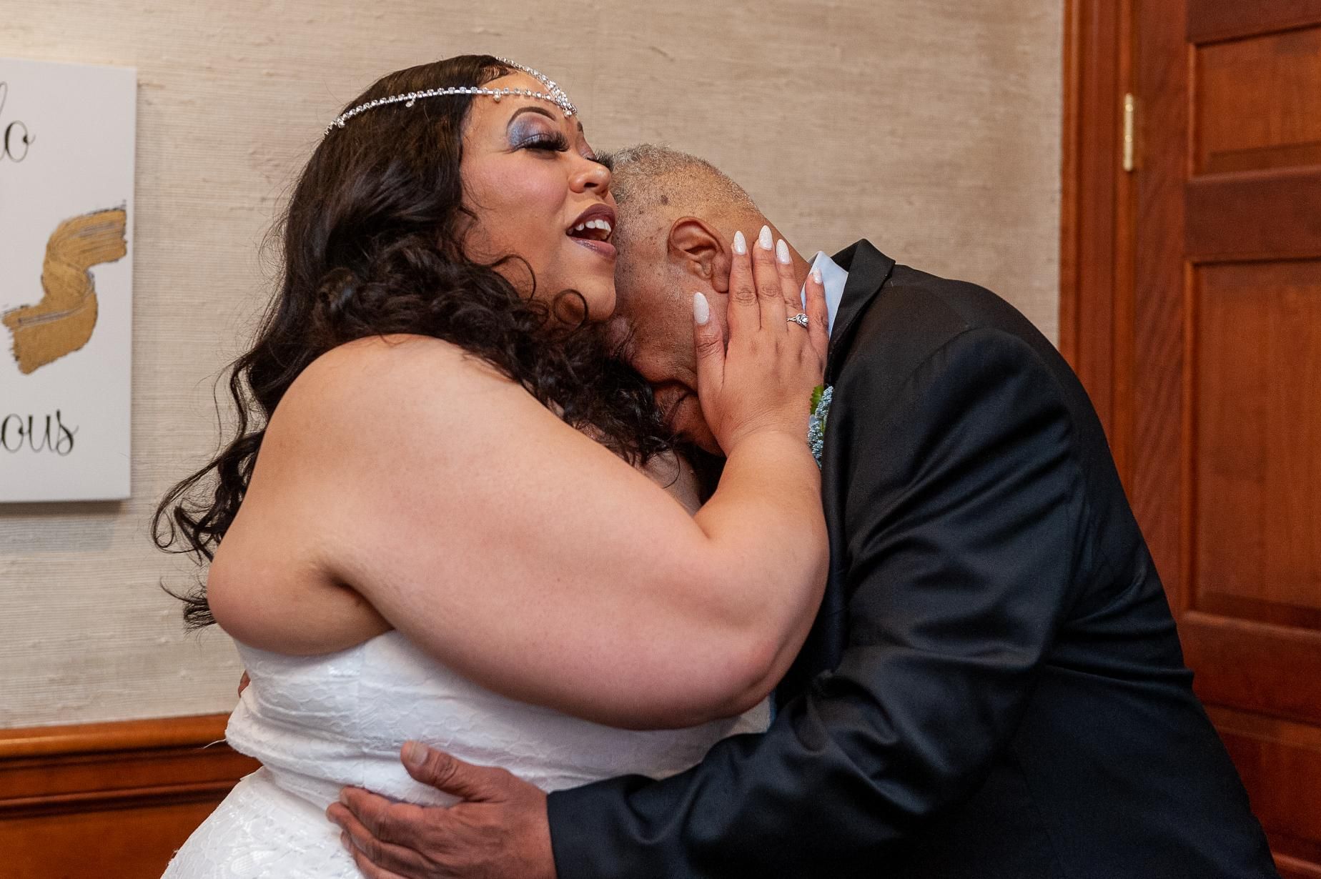 Bride in white dress hugs and kisses older man in a suit; joyful expressions in an interior setting.