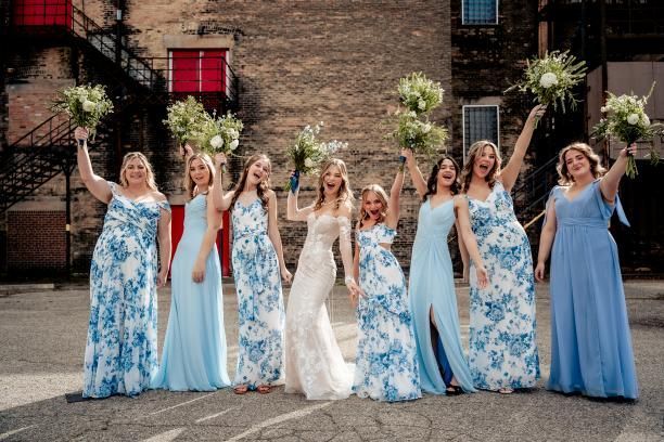 Bride and bridesmaids in blue floral and solid gowns pose outdoors with bouquets.
