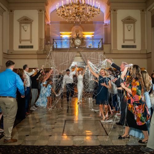 Newlyweds walk through a confetti-filled aisle as guests celebrate in a grand hall.