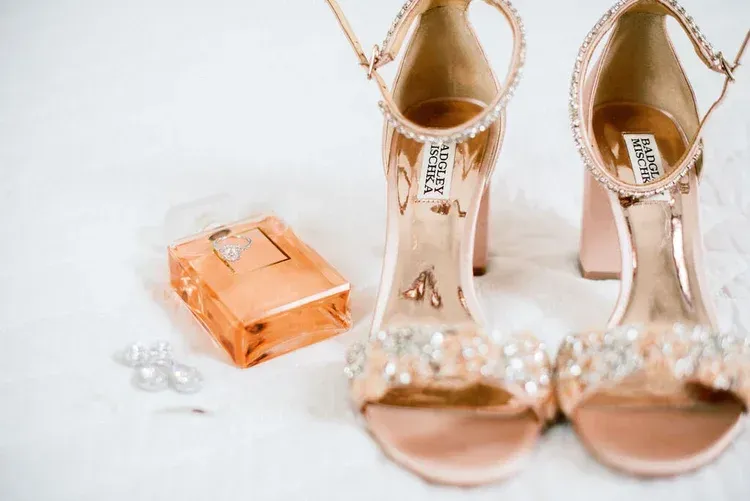Rose-gold wedding accessories: high heels, perfume, and earrings sit on a white surface.