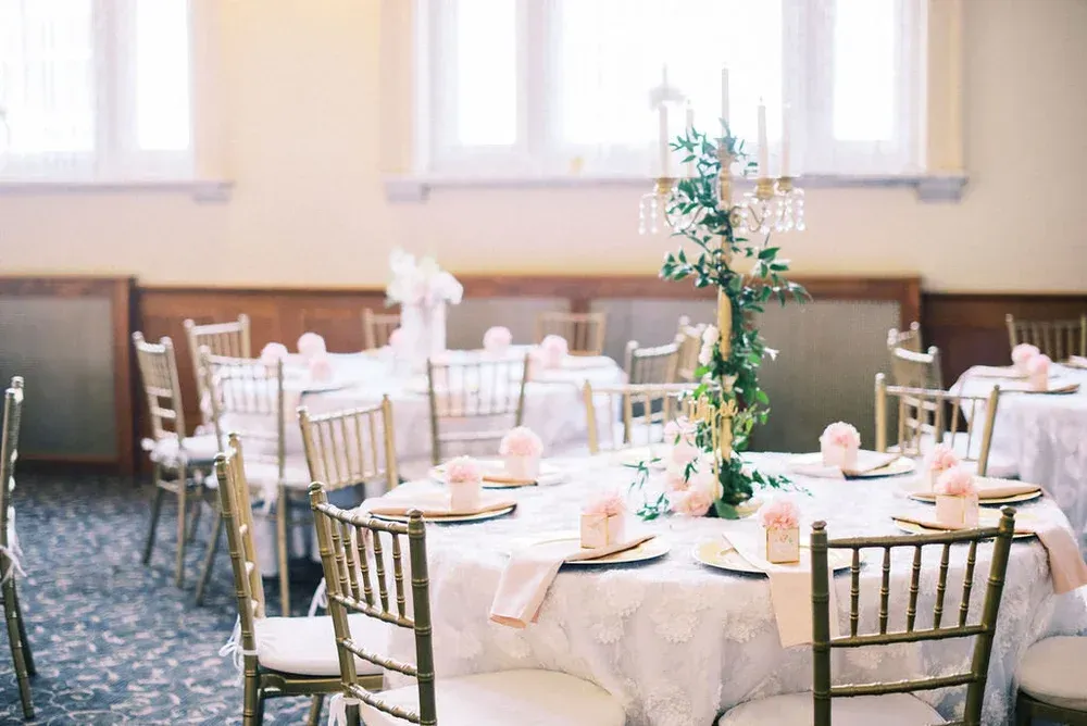 Elegant banquet tables set for a wedding, with gold chairs, white tablecloths, pink napkins, and floral centerpieces.