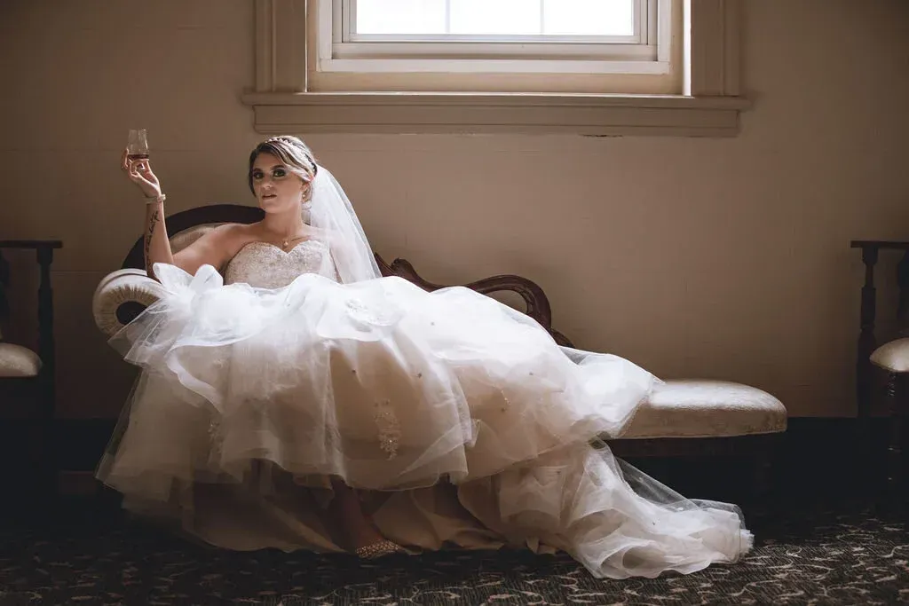 Bride in white gown reclines on chaise lounge, holding champagne. Natural light streams through a window in the background.