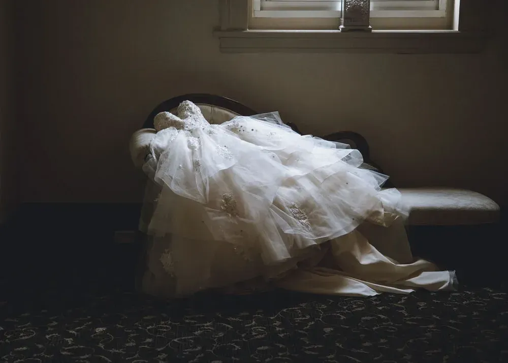 Wedding dress draped over a chaise lounge in a dimly lit room. The gown is white with layered tulle and embellishments.