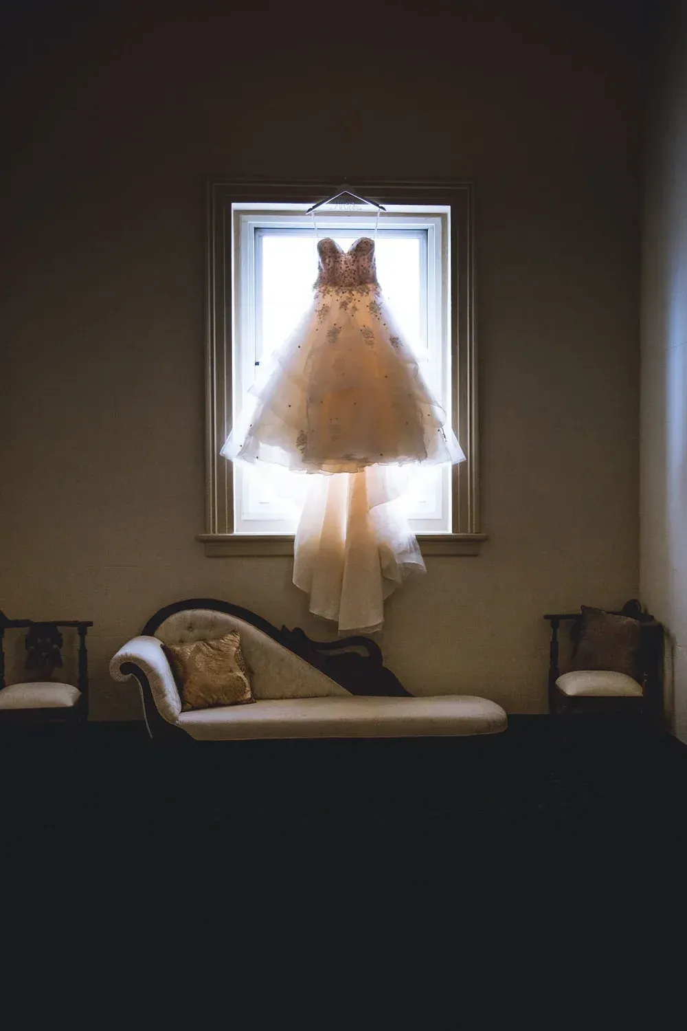 Wedding dress hanging in a window, casting a glow on the couch and chairs below in a dimly lit room.