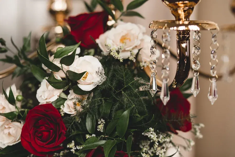 Close-up of a floral arrangement featuring red and white roses, green foliage, and a gold candelabra with hanging crystals.