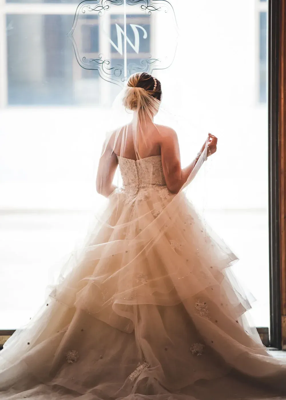 Bride in a tiered wedding dress, facing away from the camera, looking out a window.