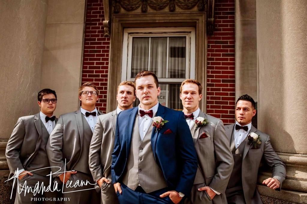 Groomsmen in suits, standing outside a building. One in a blue suit, others gray, all with bow ties.